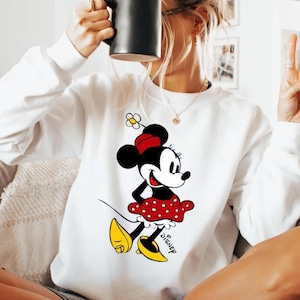 Puede incluir: Sudadera blanca con un gr&aacute;fico de Minnie Mouse. El personaje de dibujos animados lleva una falda roja de lunares, zapatos amarillos y un lazo rojo. El logotipo de Disney est&aacute; impreso debajo. La sudadera tiene mangas largas.