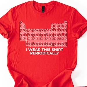 May include: A red t-shirt featuring a white periodic table of elements design. The text "I WEAR THIS SHIRT PERIODICALLY" is printed below the table. The shirt is made of a soft material and is a science-themed apparel.