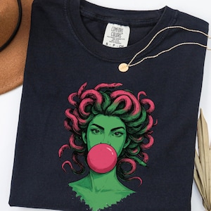 Può includere: Una maglietta blu navy con una grafica di una donna con la pelle verde, i capelli rosa e una bolla di gomma da masticare rosa. La maglietta ha un'etichetta Comfort Colors ed è abbinata a una collana d'oro.
