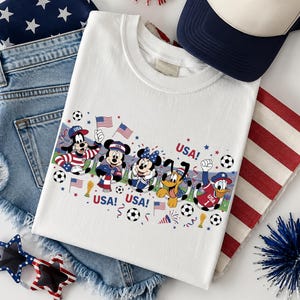 Pu&ograve; includere: Maglietta bianca con personaggi Disney in abiti patriottici, la parola "USA" e palloni da calcio. L'immagine include anche pantaloncini di jeans, un cappellino da camionista blu navy e bianco e occhiali da sole a forma di stella.