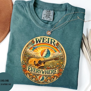 May include: A blue-green t-shirt with a circular graphic featuring a guitar, rainbow, and the words "WEIR EVERYWHERE." The shirt has a crew neck and short sleeves. A gold necklace is draped across the shirt.