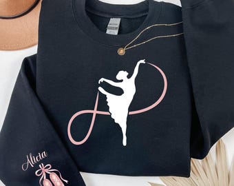 Könnte beinhalten: Schwarzes Sweatshirt mit einer weißen Ballerina-Grafik und einem rosa Band-Design. Der linke Ärmel zeigt den Namen "Alicia" in Schreibschrift sowie eine Illustration von rosa Ballettschuhen. Eine goldene Halskette und ein brauner Hut sind ebenfalls sichtbar.