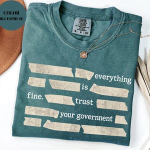 Könnte beinhalten: Ein blaugrünes T-Shirt mit dem weißen Text "everything is fine. trust your government". Der Text ist so angeordnet, als wäre er auf das T-Shirt geklebt. Eine goldene Halskette mit Anhänger liegt auf dem T-Shirt.