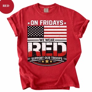 Może przedstawiać: Czerwona koszulka z podwiniętymi rękawami. Koszulka przedstawia grafikę amerykańskiej flagi i napisy "ON FRIDAYS WE WEAR RED SUPPORT OUR TROOPS" w kolorze białym i czerwonym. Czerwone koło ze słowem "RED" znajduje się w lewym górnym rogu.