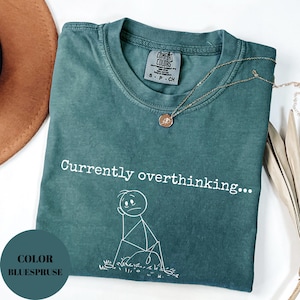 K&ouml;nnte beinhalten: Ein teal Comfort Colors T-Shirt mit dem Text "Currently overthinking..." und einer Strichm&auml;nnchen-Grafik. Eine goldene Halskette mit Anh&auml;nger liegt auf dem Shirt. Das Shirt ist gefaltet und mit einem braunen Hut ausgestellt.