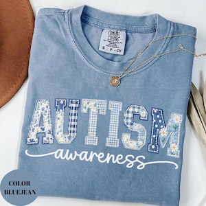 May include: Bluejean-colored t-shirt with the word "AUTISM" in patchwork letters and the word "awareness" in script. The shirt has a crew neck and a small tag on the upper back. A gold necklace with a pendant is draped across the shirt.