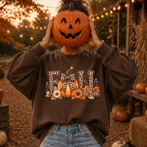 Könnte beinhalten: Ein dunkelbraunes Langarm-Sweatshirt mit dem Wort FALL in Leopardenmuster-Optik, einem zentralen Kürbis-Motiv mit Leoparden-Schleife und Sonnenblumen, kombiniert mit einer hellblauen Jeans mit leichten Abnutzungserscheinungen.