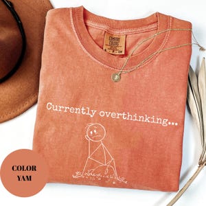 K&ouml;nnte beinhalten: Korallfarbenes T-Shirt mit dem Schriftzug "Currently overthinking..." und einer Strichm&auml;nnchen-Grafik. Das Shirt hat einen Rundhalsausschnitt und ein kleines Etikett. Eine goldene Halskette mit Anh&auml;nger liegt auf dem Shirt.