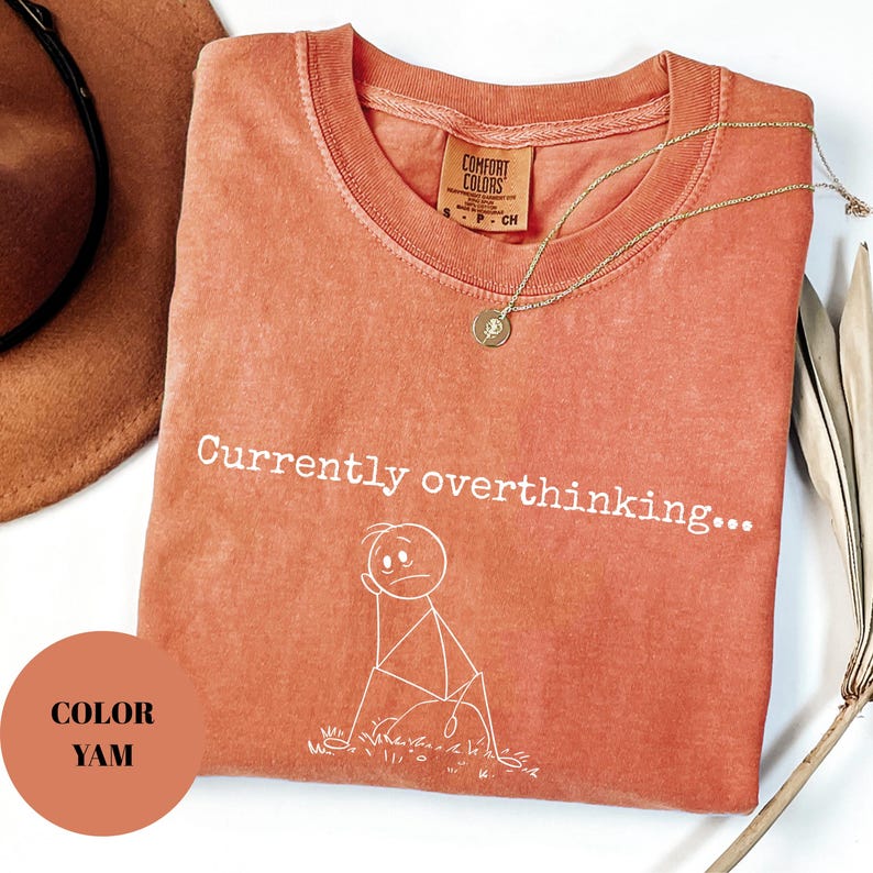 K&ouml;nnte beinhalten: Korallfarbenes T-Shirt mit dem Schriftzug "Currently overthinking..." und einer Strichm&auml;nnchen-Grafik. Das Shirt hat einen Rundhalsausschnitt und ein kleines Etikett. Eine goldene Halskette mit Anh&auml;nger liegt auf dem Shirt.