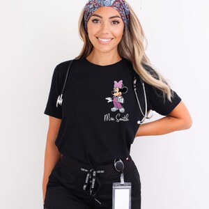 以下が含まれることがあります： ピンクと紫のミニーマウスのグラフィックが特徴の黒いTシャツ。「Mrs. Smith」という名前が白い文字でプリントされています。聴診器が見えます。モデルは模様入りのヘッドバンドを着用しています。