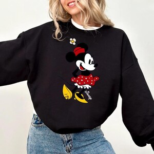 Puede incluir: Sudadera negra con un gr&aacute;fico de Minnie Mouse. El gr&aacute;fico muestra a Minnie Mouse con una falda roja de lunares, un lazo rojo y zapatos amarillos. La palabra "Disney" est&aacute; impresa debajo.