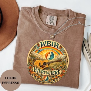 May include: Espresso-coloured t-shirt with a circular graphic featuring a guitar, rainbow, and the words "WEIR EVERYWHERE." The design includes flowers, mushrooms, and a stylized sun. The shirt has a crew neck and short sleeves. A silver necklace with a pendant is also visible.