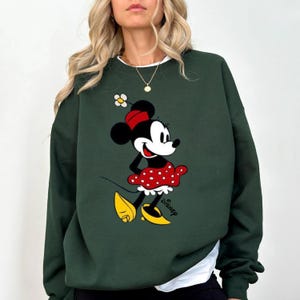 Puede incluir: Sudadera verde oscuro con un gr&aacute;fico de Minnie Mouse. El personaje de dibujos animados lleva una falda roja de lunares, un lazo rojo y zapatos amarillos. La palabra "Disney" est&aacute; impresa debajo.