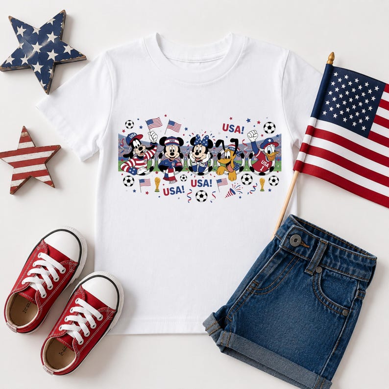 Pu&ograve; includere: Maglietta bianca con personaggi Disney in abiti patriottici, la scritta "USA!" e palloni da calcio. Scarpe da ginnastica rosse e pantaloncini di jeans sono accanto alla maglietta, insieme a una piccola bandiera americana e decorazioni a forma di stella.