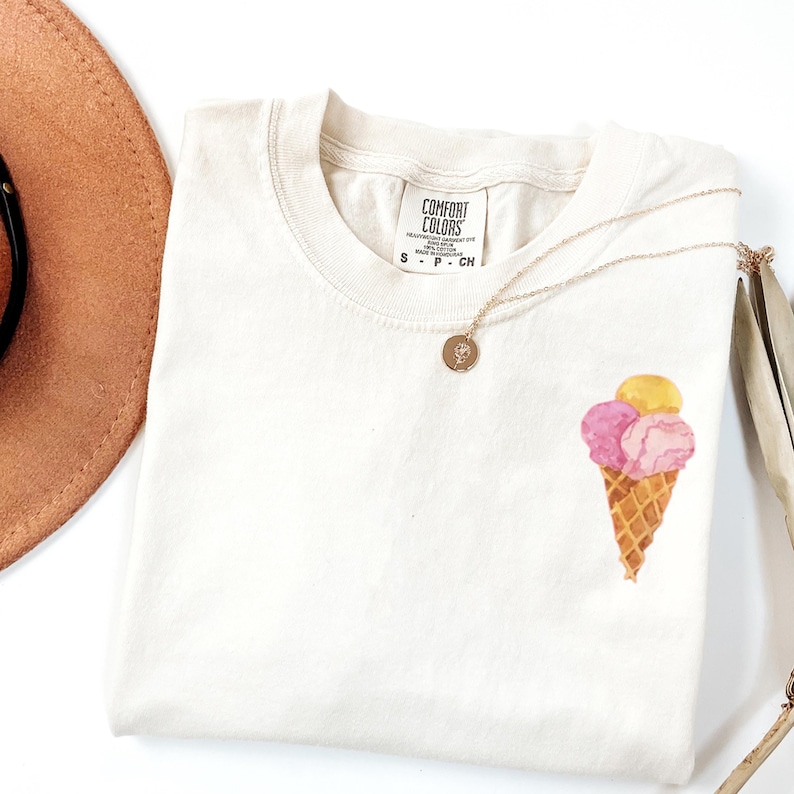 May include: Cream-colored t-shirt with a watercolor ice cream cone design. The cone is brown, and the scoops are pink and yellow. A gold necklace with a pendant rests on the shirt. The shirt has a tag that says "Comfort Colors".