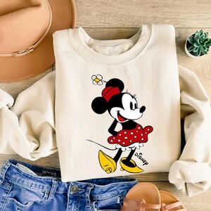 Puede incluir: Sudadera de color crema con un gr&aacute;fico de Minnie Mouse. Minnie est&aacute; representada con una falda roja de lunares, zapatos amarillos y un lazo rojo. El logotipo de Disney est&aacute; impreso en la parte inferior derecha. La sudadera se combina con vaqueros azules y un sombrero marr&oacute;n.