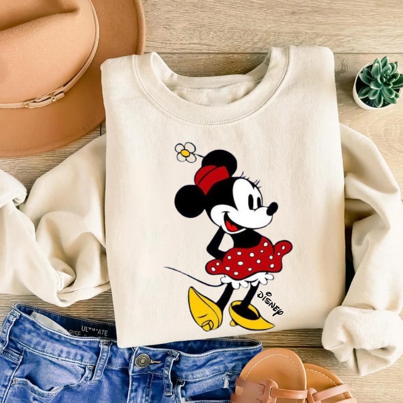 Puede incluir: Sudadera de color crema con un gr&aacute;fico de Minnie Mouse. Minnie est&aacute; representada con una falda roja de lunares, zapatos amarillos y un lazo rojo. El logotipo de Disney est&aacute; impreso en la parte inferior derecha. La sudadera se combina con vaqueros azules y un sombrero marr&oacute;n.