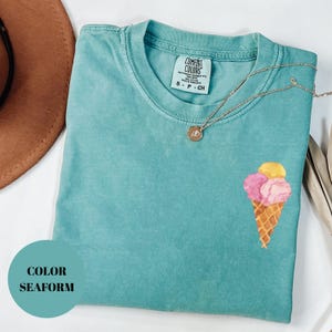 May include: Seafoam green t-shirt with an embroidered ice cream cone design. The shirt has a crew neck and short sleeves. A gold necklace with a pendant is draped across the shirt. The tag reads "Comfort Colors".