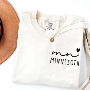 K&ouml;nnte beinhalten: Cremefarbenes T-Shirt mit dem Text "mn MINNESOTA" in schwarzer Schrift und einem kleinen schwarzen Herz. Das Shirt hat einen runden Ausschnitt und ein Etikett mit der Aufschrift "Comfort Colors". Eine goldene Halskette mit Anh&auml;nger liegt &uuml;ber dem Shirt.