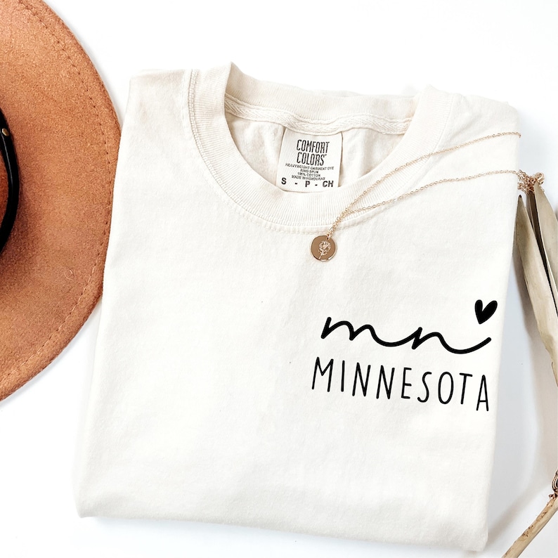 K&ouml;nnte beinhalten: Cremefarbenes T-Shirt mit dem Text "mn MINNESOTA" in schwarzer Schrift und einem kleinen schwarzen Herz. Das Shirt hat einen runden Ausschnitt und ein Etikett mit der Aufschrift "Comfort Colors". Eine goldene Halskette mit Anh&auml;nger liegt &uuml;ber dem Shirt.