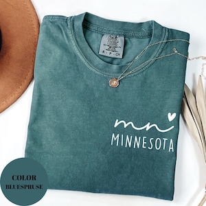 K&ouml;nnte beinhalten: Ein teal-farbenes Comfort Colors T-Shirt mit dem Text "mn" und "MINNESOTA" in wei&szlig;er Schrift. Eine goldene Halskette mit Anh&auml;nger liegt auf dem Shirt. Das Shirt ist gefaltet, und ein brauner Hut ist im Hintergrund zu sehen.