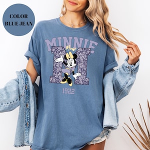 Pu&ograve; includere: T-shirt blu jeans con Minnie Mouse. Il design include la parola "MINNIE" in lavanda, il personaggio Minnie Mouse e l'anno "1922". La lettera "M" &egrave; riempita con un motivo floreale.