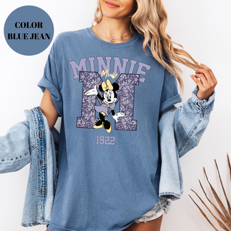 Pu&ograve; includere: T-shirt blu jeans con Minnie Mouse. Il design include la parola "MINNIE" in lavanda, il personaggio Minnie Mouse e l'anno "1922". La lettera "M" &egrave; riempita con un motivo floreale.