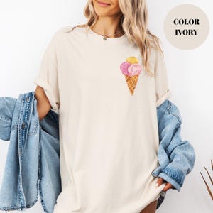 May include: Ivory-colored t-shirt featuring a watercolor ice cream cone design with pink and yellow scoops. The shirt has a relaxed fit and rolled-up sleeves. The text "COLOR IVORY" is in a cream-colored circle.