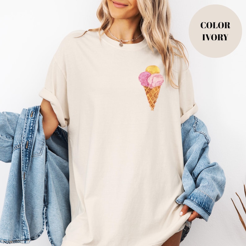 May include: Ivory-colored t-shirt featuring a watercolor ice cream cone design with pink and yellow scoops. The shirt has a relaxed fit and rolled-up sleeves. The text "COLOR IVORY" is in a cream-colored circle.