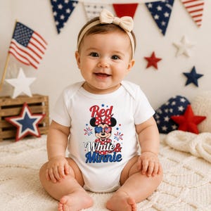 K&ouml;nnte beinhalten: Ein Baby tr&auml;gt einen wei&szlig;en Body mit einem Minnie-Maus-Motiv in Rot, Wei&szlig; und Blau. Der Aufdruck lautet "Red, White & Minnie". Das Baby tr&auml;gt ein beiges Schleifen-Stirnband und l&auml;chelt. Amerikanische Flaggen und Stern-Dekorationen sind im Hintergrund.