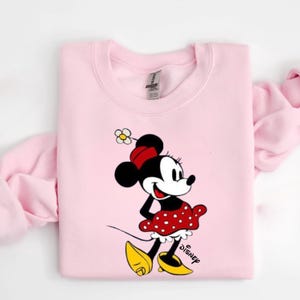 Puede incluir: Sudadera rosa claro con un dise&ntilde;o de Minnie Mouse. Minnie lleva su atuendo cl&aacute;sico: una falda roja de lunares, zapatos amarillos y un lazo rojo. La palabra "Disney" est&aacute; impresa en negro debajo.