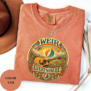 May include: A yam-coloured t-shirt with a round graphic featuring a guitar, rainbow, and the words "WEIR EVERYWHERE." The shirt has a crew neck and a small tag. A gold necklace is draped across the shirt.