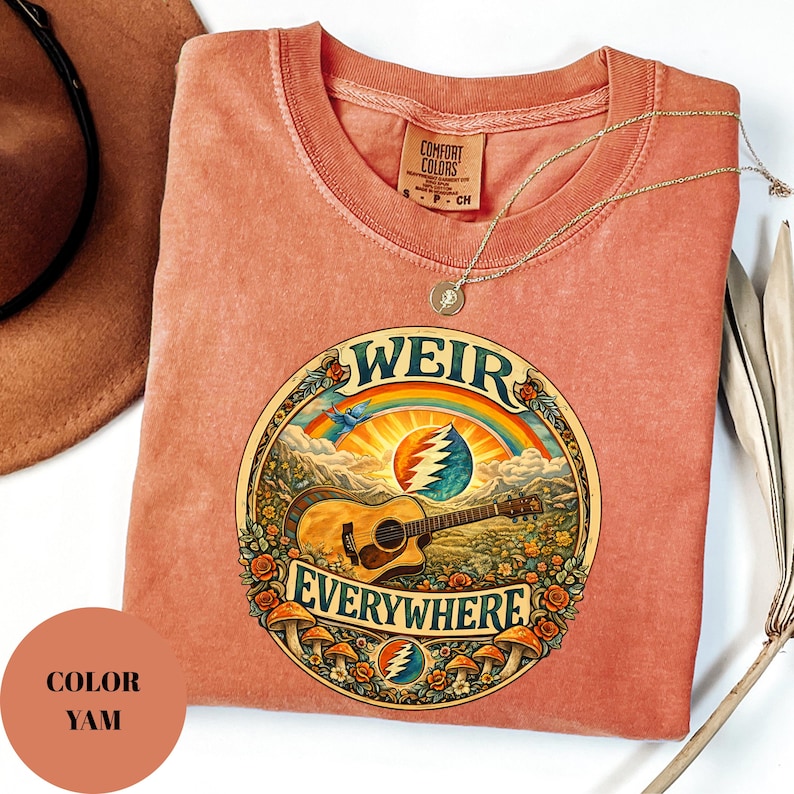 May include: A yam-coloured t-shirt with a round graphic featuring a guitar, rainbow, and the words "WEIR EVERYWHERE." The shirt has a crew neck and a small tag. A gold necklace is draped across the shirt.