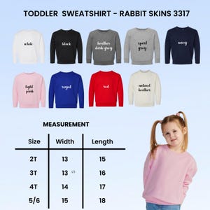 Puede incluir: Una selecci&oacute;n de sudaderas para ni&ntilde;os peque&ntilde;os en varios colores, incluyendo blanco, negro y azul marino. La imagen muestra las sudaderas con el texto "TODDLER SWEATSHIRT - RABBIT SKINS 3317". Una tabla de medidas muestra tallas 2T a 5/6 con ancho y largo en cm.