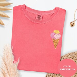May include: A coral-colored t-shirt with a watercolor ice cream cone design. The ice cream cone has three scoops in yellow, pink, and purple. The shirt has a crew neck and the brand "Comfort Colors" is visible on the tag.