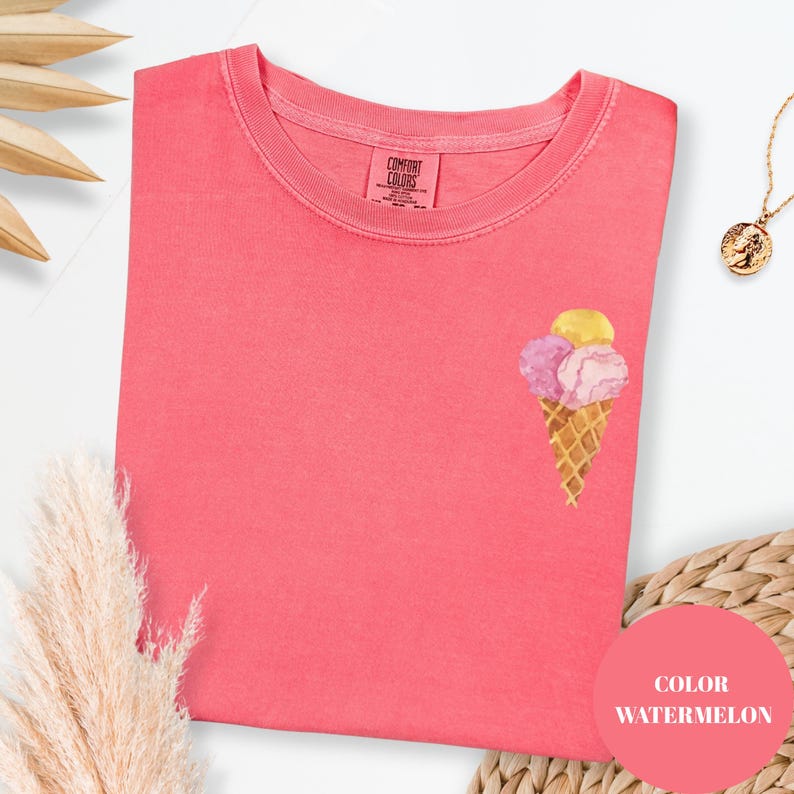 May include: A coral-colored t-shirt with a watercolor ice cream cone design. The ice cream cone has three scoops in yellow, pink, and purple. The shirt has a crew neck and the brand "Comfort Colors" is visible on the tag.