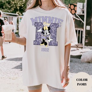 Pu&ograve; includere: T-shirt color avorio con grafica di Minnie Mouse. Il design include una "M" viola con motivo floreale, la scritta "MINNIE" e il numero "1922". La maglietta ha una vestibilit&agrave; ampia.