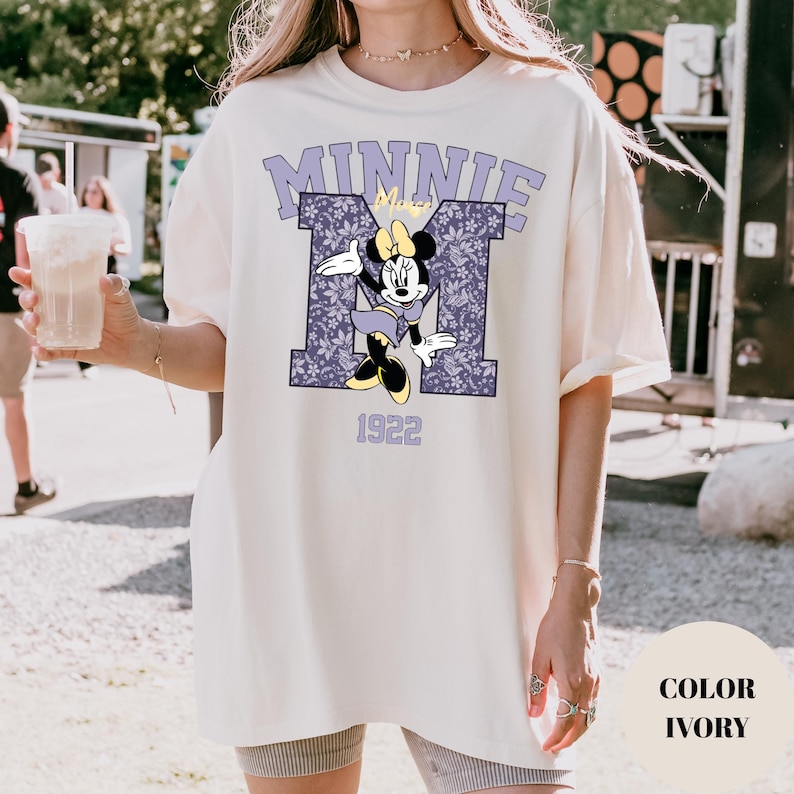 Pu&ograve; includere: T-shirt color avorio con grafica di Minnie Mouse. Il design include una "M" viola con motivo floreale, la scritta "MINNIE" e il numero "1922". La maglietta ha una vestibilit&agrave; ampia.
