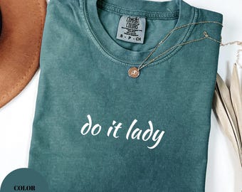 Puede incluir: Una camiseta Comfort Colors de color verde azulado con la frase "do it lady" en escritura blanca. Un collar de oro con un colgante descansa sobre la camisa. La camisa está doblada y un sombrero marrón es parcialmente visible.