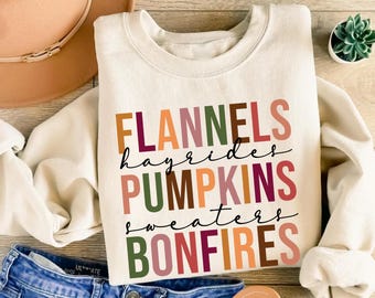 May include: A cream-coloured sweatshirt featuring the words "FLANNELS, hayrides, PUMPKINS, sweaters, BONFIRES" in autumnal hues. The sweatshirt is styled with blue jeans, a brown hat, and sandals.