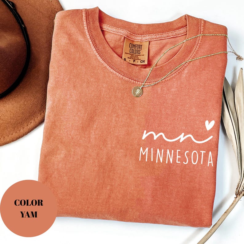 K&ouml;nnte beinhalten: Ein yam-farbenes T-Shirt mit dem wei&szlig;en Aufdruck "Minnesota". Das Shirt hat einen Rundhalsausschnitt und eine kleine Herzgrafik. Eine goldene Halskette mit Anh&auml;nger liegt &uuml;ber dem Shirt.