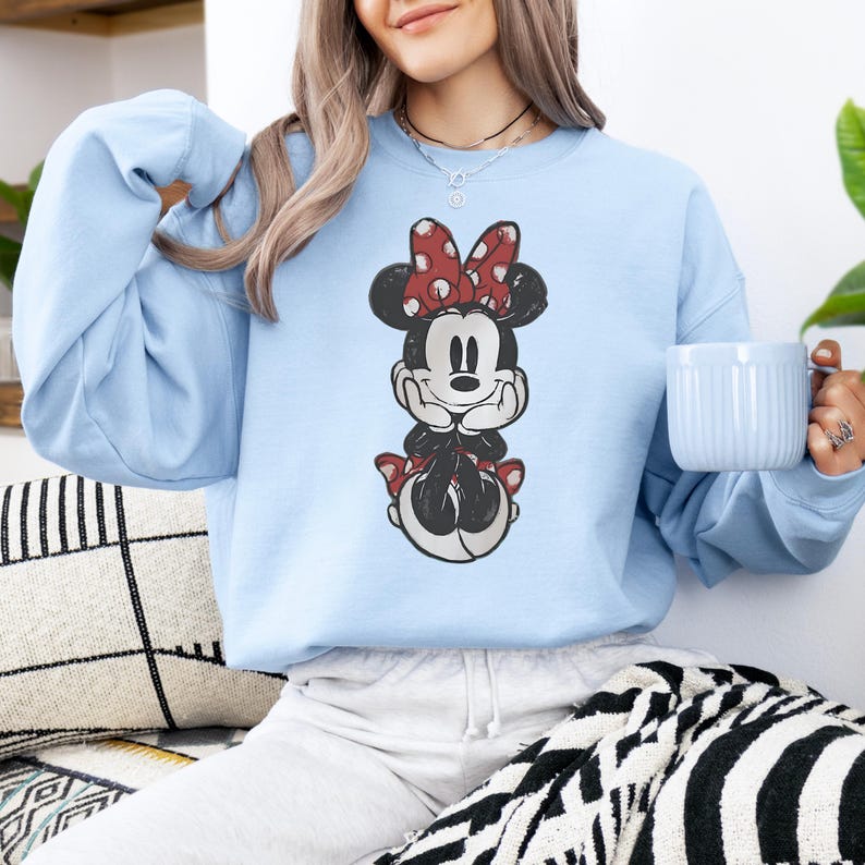 May include: Light blue sweatshirt featuring a distressed graphic of Minnie Mouse. Minnie is depicted in black, white, and red, with a red polka dot bow. The sweatshirt is a solid light blue color.