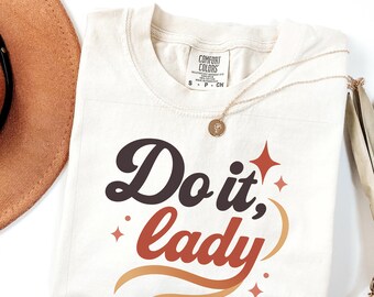 Puede incluir: Camiseta color crema con la frase "Do it, lady" en letras marrones y naranjas. La camiseta tiene un collar dorado y está junto a un sombrero marrón.