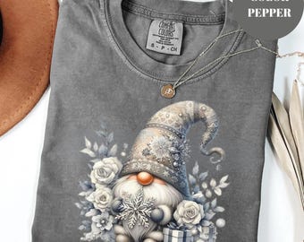 May include: A gray Comfort Colors t-shirt featuring a whimsical gnome design. The gnome wears a detailed hat and is surrounded by white roses, snowflakes, and a gift box. A silver necklace with a pendant is draped across the shirt.