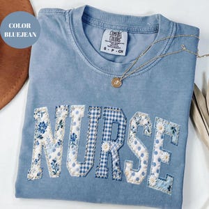 以下が含まれることがあります： ブルーのジーンズカラーのTシャツ。"NURSE"の文字が花柄、ギンガムチェック、水玉模様でアップリケされています。ペンダント付きのゴールドネックレスがシャツの上にあります。シャツには「Comfort Colors」のラベルがあり、サイズはSです。