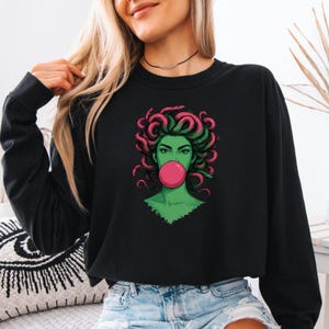 May include: Black long-sleeve sweatshirt featuring a graphic of a green-skinned woman with pink and green hair blowing a pink bubble. The graphic is a stylized illustration of Medusa.