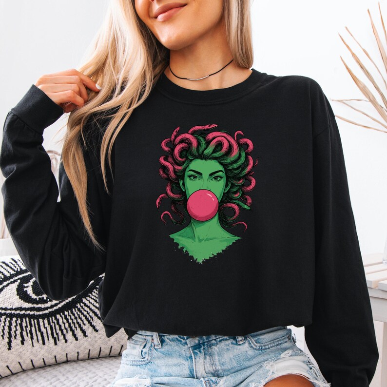 May include: Black long-sleeve sweatshirt featuring a graphic of a green-skinned woman with pink and green hair blowing a pink bubble. The graphic is a stylized illustration of Medusa.