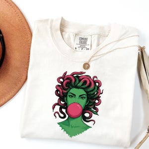 May include: Cream-colored t-shirt featuring a graphic of a green-skinned woman with pink Medusa-like hair blowing a pink bubble. The shirt has a Comfort Colors tag and a gold necklace. The shirt is folded.