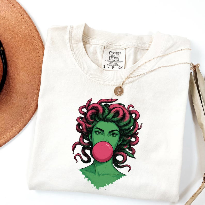 May include: Cream-colored t-shirt featuring a graphic of a green-skinned woman with pink Medusa-like hair blowing a pink bubble. The shirt has a Comfort Colors tag and a gold necklace. The shirt is folded.