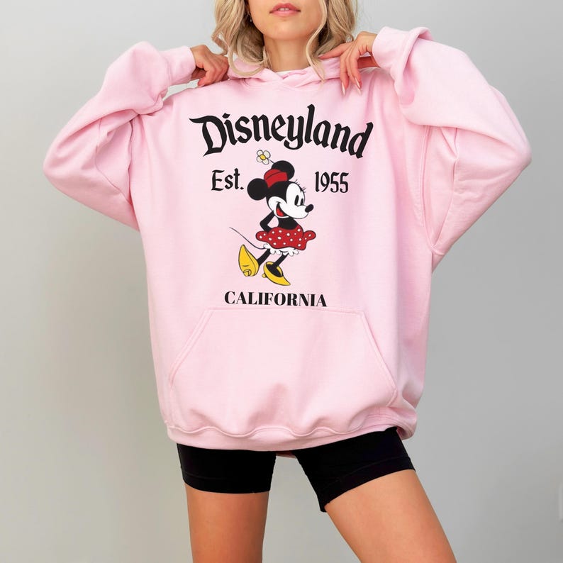 May include: A pale pink Disneyland hoodie with a Minnie Mouse design. The hoodie has a front pocket and the words "Disneyland Est. 1955 CALIFORNIA" printed in black. The graphic shows Minnie Mouse in a red polka dot skirt.